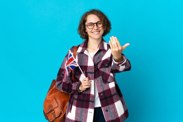 Jovem mulher inglesa segurando uma bandeira do Reino Unido isolada na parede azul, convidando para vir com a mão. Feliz que você veio