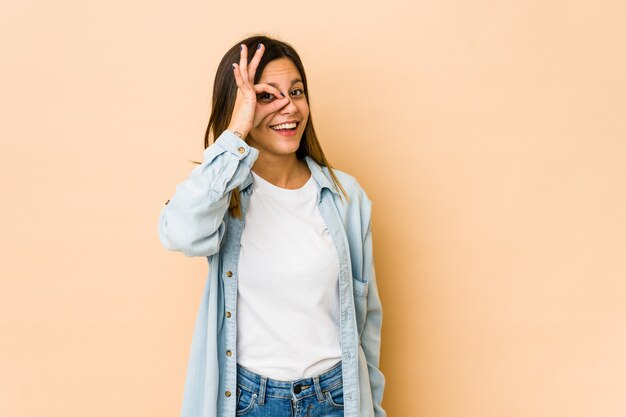 Jovem mulher isolada no espaço bege animado mantendo o gesto ok no olho.