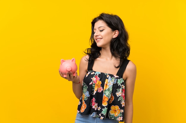 Jovem mulher isolada parede amarela segurando um grande piggybank