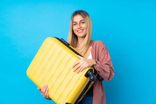 Jovem mulher isolada parede azul em férias com mala de viagem e um chapéu