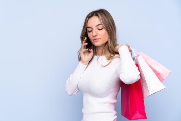 Jovem mulher isolada parede azul segurando sacolas de compras e chamando um amigo com seu telefone celular