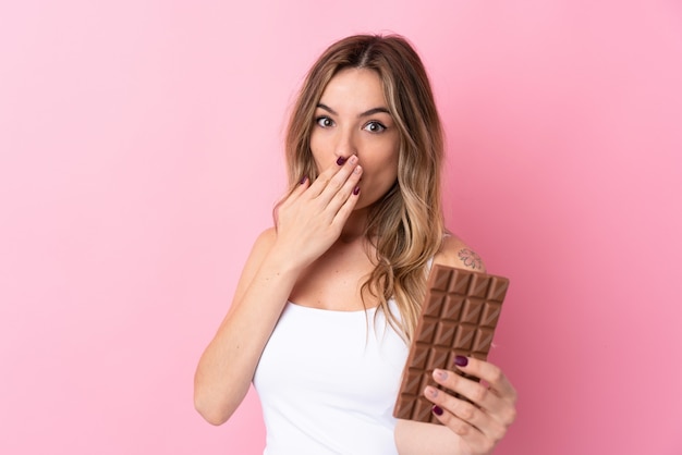 Foto jovem mulher isolada parede rosa tomando uma tablete de chocolate e surpreendeu