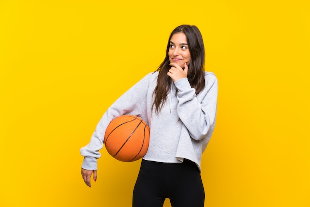 Jovem mulher jogando basquete sobre parede amarela isolada, pensando uma idéia