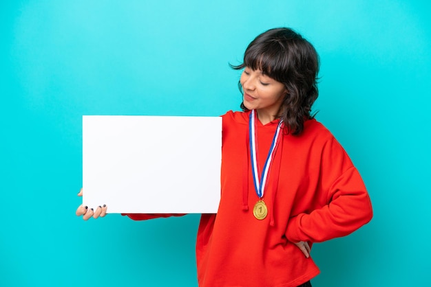 Jovem mulher latina com medalhas isoladas em fundo azul segurando um cartaz vazio com expressão feliz e apontando-o