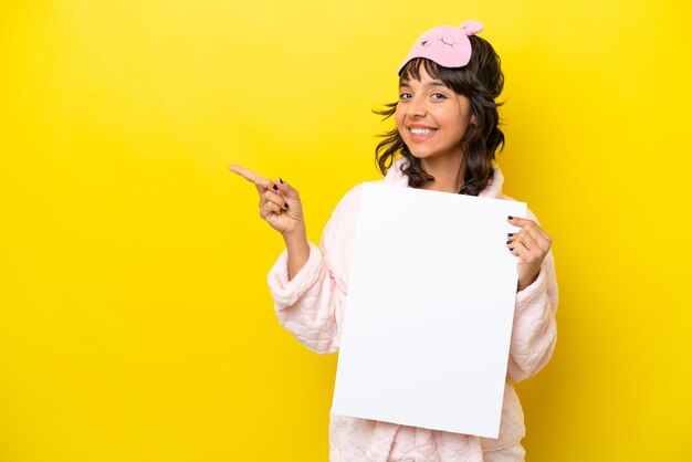 Jovem mulher latina de pijama isolada em fundo amarelo segurando um cartaz vazio e apontando o lado