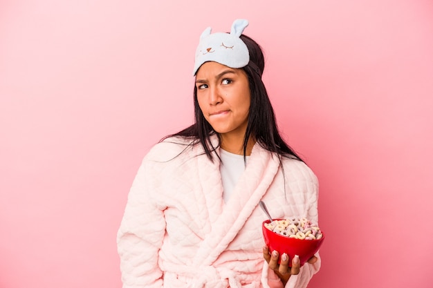 Jovem mulher latina de pijama segurando uma tigela de cereais isolada no fundo rosa confusa, sente-se em dúvida e insegura.