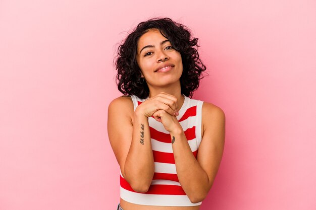 Jovem mulher latina encaracolada isolada em fundo rosa mantém as mãos sob o queixo, olhando alegremente de lado.