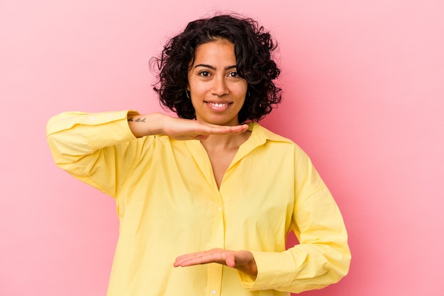 Jovem mulher latina encaracolada isolada no fundo rosa, segurando algo com as duas mãos, apresentação do produto.