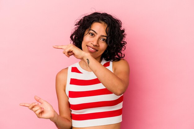 Jovem mulher latina encaracolada isolada no fundo rosa, segurando algo pequeno com os indicadores, sorrindo e confiante.