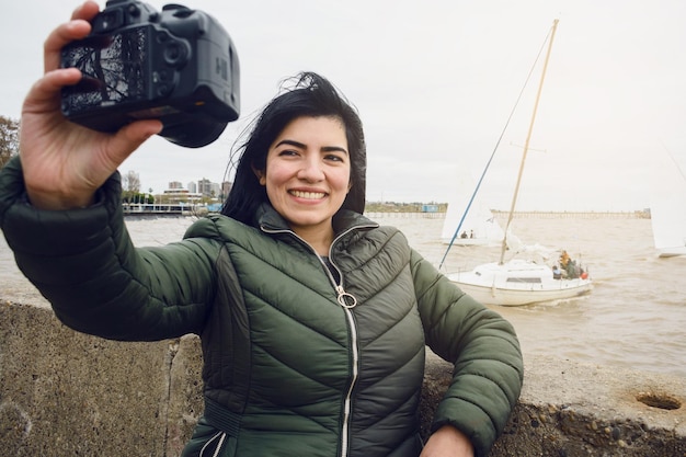jovem mulher latina feliz tirando uma selfie com sua câmera com o rio e barcos no fundo