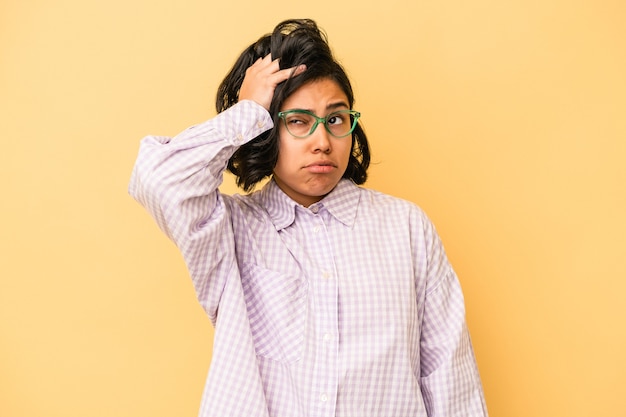 Jovem mulher latina isolada em fundo amarelo, cansada e com muito sono, mantendo a mão na cabeça.