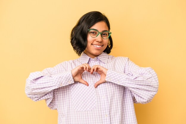 Jovem mulher latina isolada em fundo amarelo, sorrindo e mostrando uma forma de coração com as mãos.