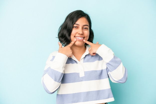 Jovem mulher latina isolada em sorrisos de fundo azul, apontando os dedos para a boca.