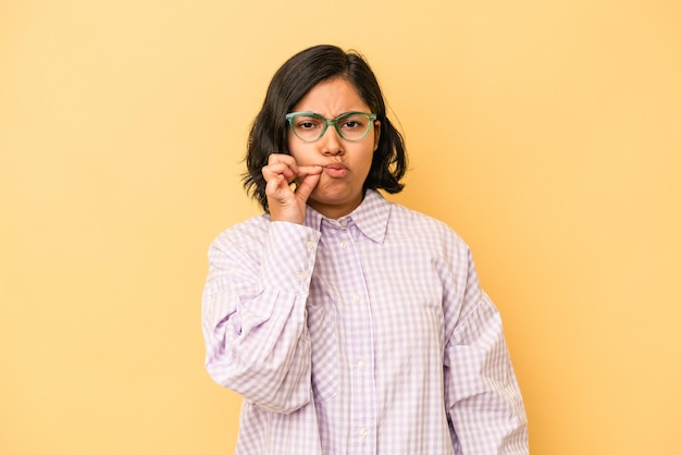 Jovem mulher latina isolada em um fundo amarelo com os dedos nos lábios, mantendo um segredo.