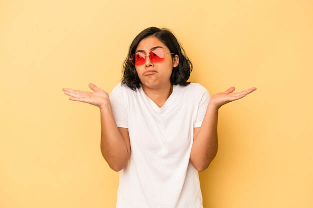 Jovem mulher latina isolada em um fundo amarelo confusa e duvidosa, encolhendo os ombros para segurar um espaço de cópia.