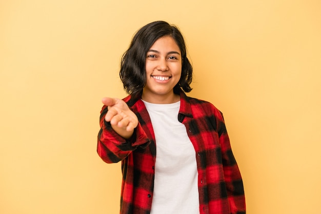 Jovem mulher latina isolada em um fundo amarelo, esticando a mão para a câmera em um gesto de saudação.