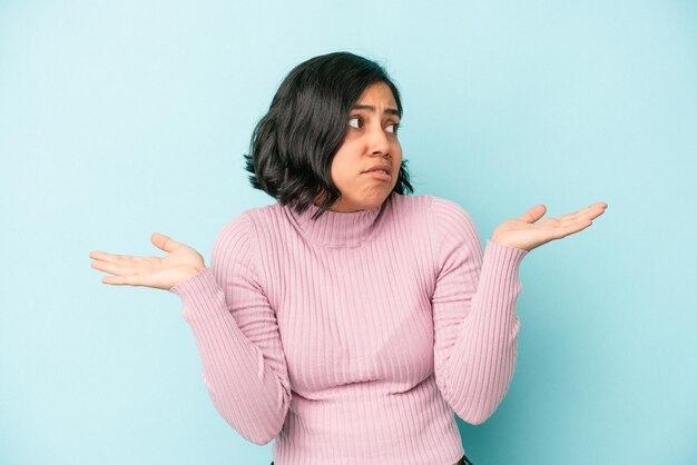 Jovem mulher latina isolada em um fundo azul confusa e duvidosa, encolhendo os ombros para segurar um espaço de cópia.