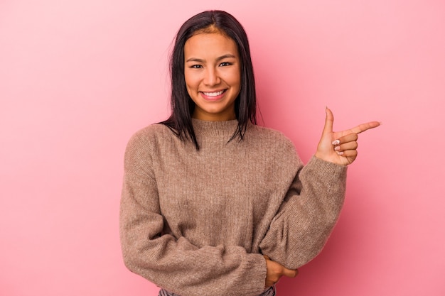 Jovem mulher latina isolada no fundo rosa, sorrindo alegremente, apontando com o dedo indicador para longe.