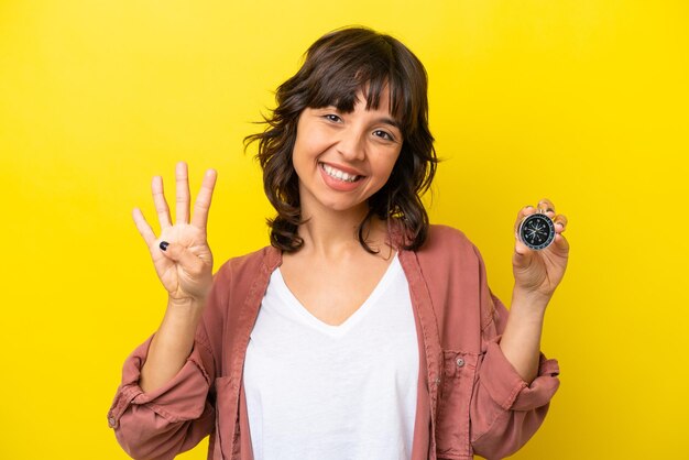 Jovem mulher latina segurando bússola isolada em fundo amarelo feliz e contando quatro com os dedos