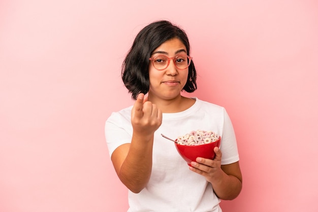 Jovem mulher latina segurando cereais isolados no fundo rosa, apontando com o dedo para você como se fosse um convite para se aproximar.