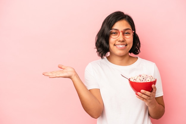 Jovem mulher latina segurando cereais isolados no fundo rosa, mostrando um espaço de cópia na palma da mão e segurando a outra mão na cintura.