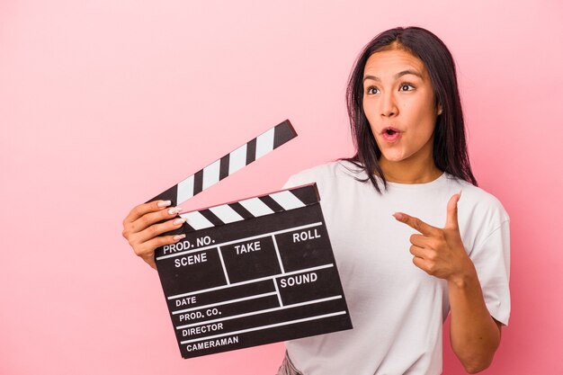 Jovem mulher latina segurando claquete isolada em um fundo rosa apontando para o lado