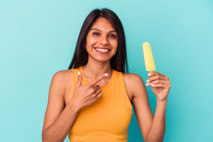 Foto jovem mulher latina segurando sorvete isolado em um fundo azul, sorrindo e apontando de lado, mostrando algo no espaço em branco.