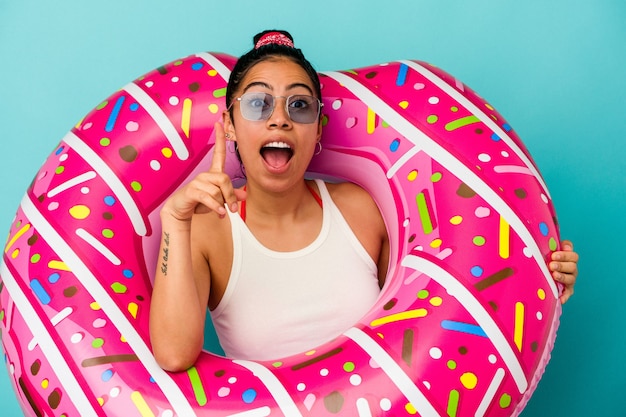 Jovem mulher latina segurando um donut inflável isolado em um fundo azul apontando de cabeça para baixo com a boca aberta.