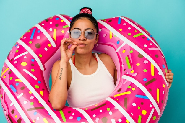 Jovem mulher latina segurando um donut inflável isolado em um fundo azul com os dedos nos lábios, mantendo um segredo.