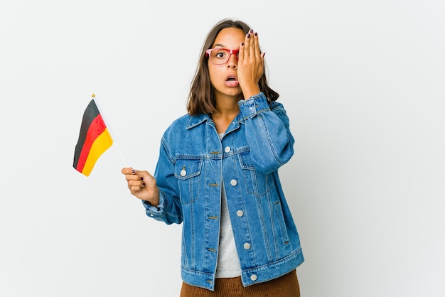 Jovem mulher latina segurando uma bandeira alemã isolada na parede branca, se divertindo cobrindo metade do rosto com a palma da mão.