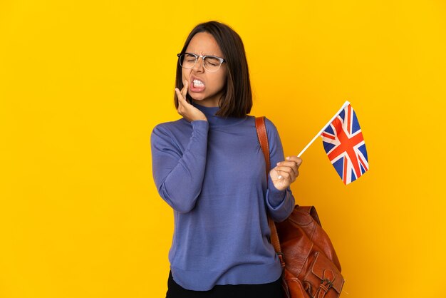 Jovem mulher latina segurando uma bandeira do Reino Unido, isolada em um fundo amarelo, com dor de dente