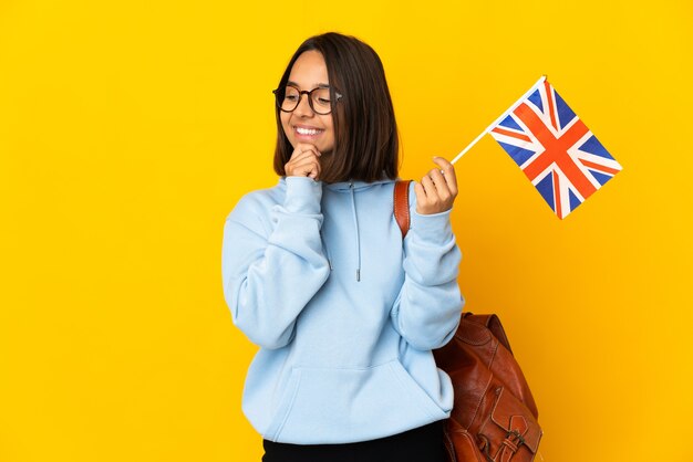 Jovem mulher latina segurando uma bandeira do Reino Unido isolada em um fundo amarelo, olhando para o lado e sorrindo