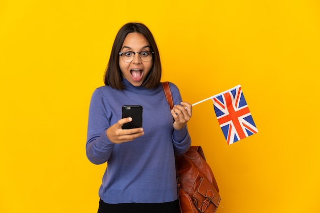 Jovem mulher latina segurando uma bandeira do reino unido isolada em um fundo amarelo surpresa e enviando uma mensagem