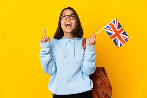 Jovem mulher latina segurando uma bandeira do Reino Unido isolada na parede amarela apontando para cima e surpresa