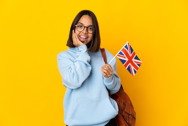 Jovem mulher latina segurando uma bandeira do Reino Unido isolada na parede amarela com expressão facial de surpresa e choque