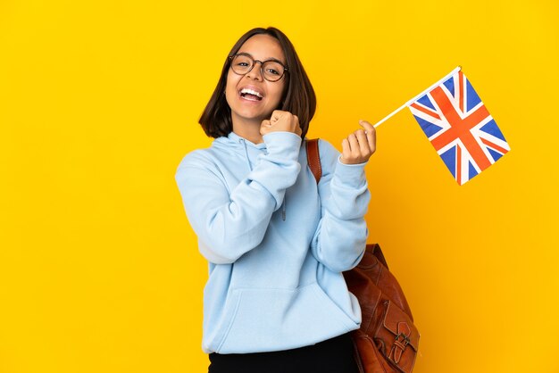 Jovem mulher latina segurando uma bandeira do Reino Unido isolada na parede amarela comemorando uma vitória