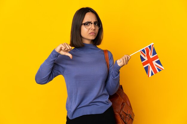 Jovem mulher latina segurando uma bandeira do Reino Unido, isolada na parede amarela, mostrando o polegar para baixo com expressão negativa