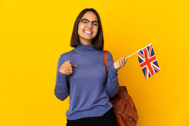 Jovem mulher latina segurando uma bandeira do Reino Unido isolada na parede amarela, orgulhosa e satisfeita
