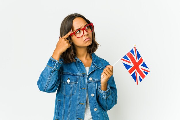 Jovem mulher latina segurando uma bandeira inglesa isolada na parede branca, apontando o templo com o dedo, pensando, focada em uma tarefa.