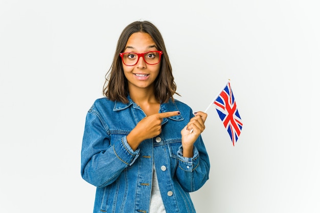 Jovem mulher latina segurando uma bandeira inglesa isolada na parede branca, sorrindo alegremente, apontando com o dedo indicador para longe.