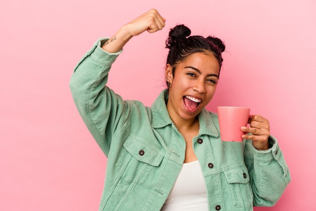 Jovem mulher latina segurando uma caneca isolada no fundo rosa, levantando o punho após uma vitória, o conceito de vencedor.