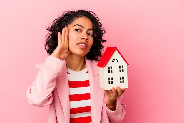 Jovem mulher latina segurando uma casa de brinquedo isolada no fundo rosa, tentando ouvir uma fofoca.