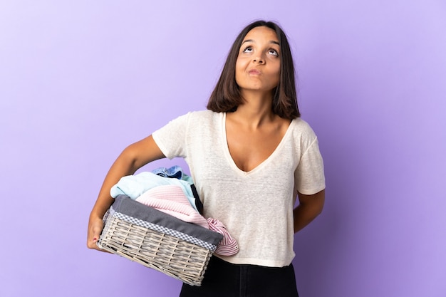 Jovem mulher latina segurando uma cesta de roupas isolada em roxo e olhando para cima