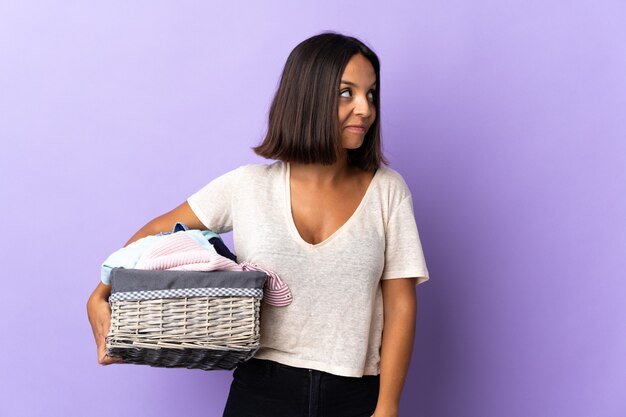 Jovem mulher latina segurando uma cesta de roupas isolada em roxo tendo dúvidas enquanto olha para cima