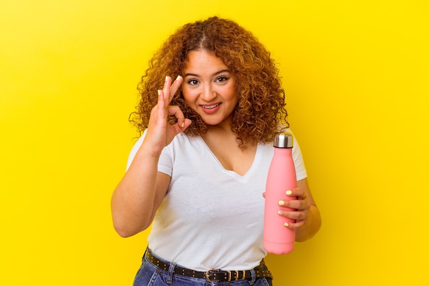 Jovem mulher latina segurando uma garrafa térmica isolada em fundo amarelo alegre e confiante, mostrando o gesto de ok.