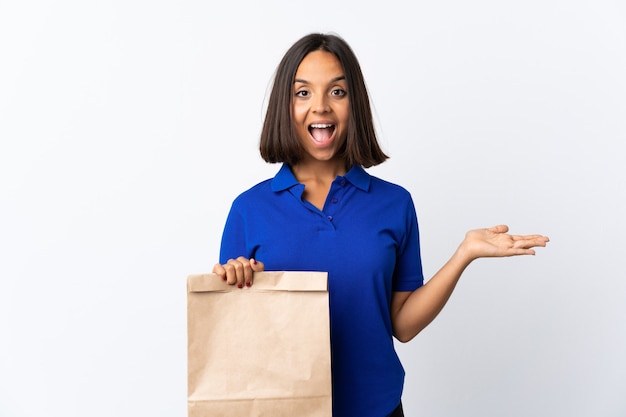 Jovem mulher latina segurando uma sacola de compras de supermercado isolada no branco com expressão facial chocada