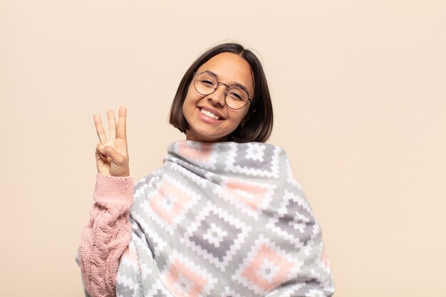 Jovem mulher latina sorrindo e parecendo amigável, mostrando o número três ou terceiro com a mão para a frente