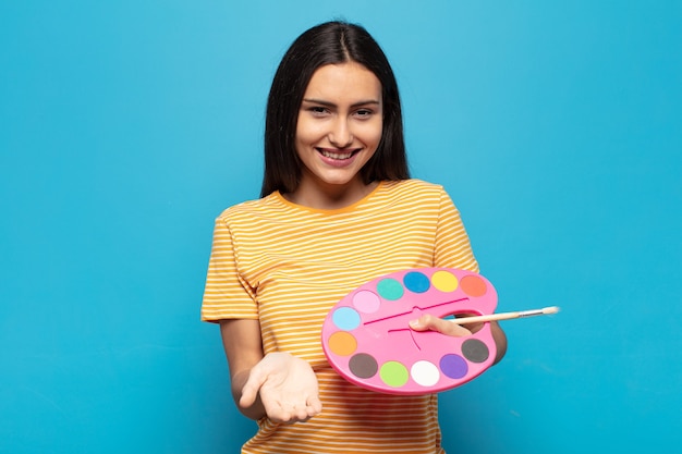 Jovem mulher latina sorrindo feliz com um olhar amigável, confiante e positivo, oferecendo e mostrando um objeto ou conceito