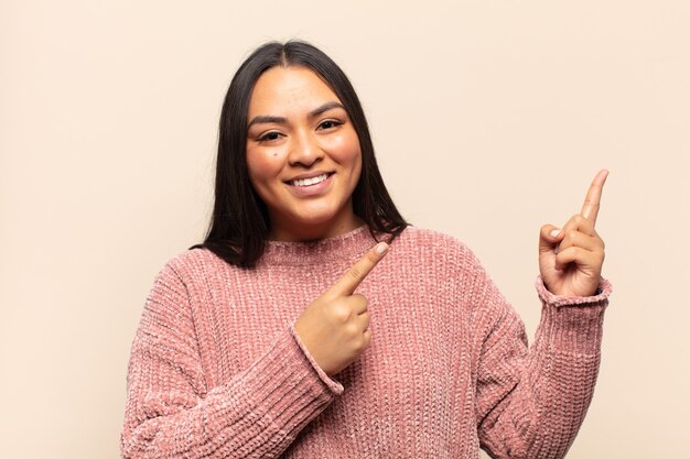 Jovem mulher latina sorrindo feliz e apontando para o lado e para cima com as duas mãos mostrando o objeto no espaço da cópia