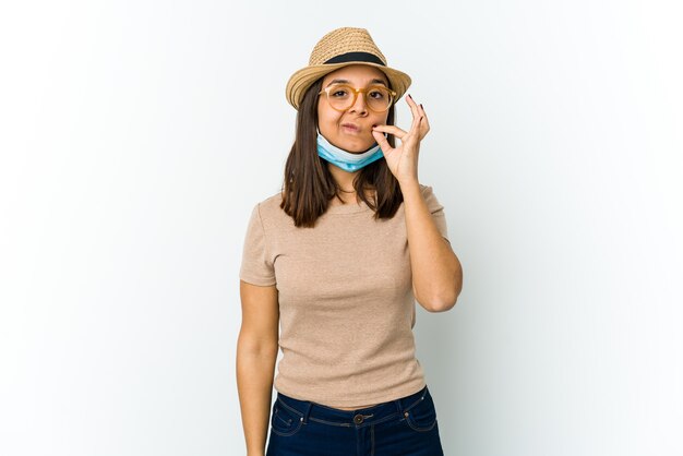 Jovem mulher latina usando chapéu e máscara para proteger de cobiçoso isolado no branco com os dedos nos lábios, mantendo um segredo.